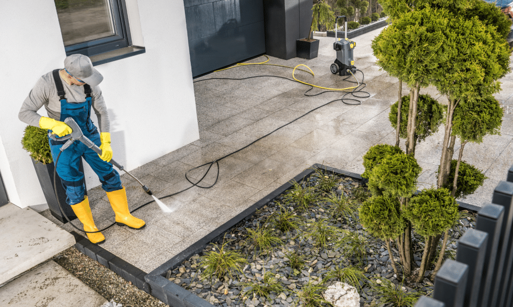 Person wearing yellow boots using a pressure washer to clean a concrete surface near a garden