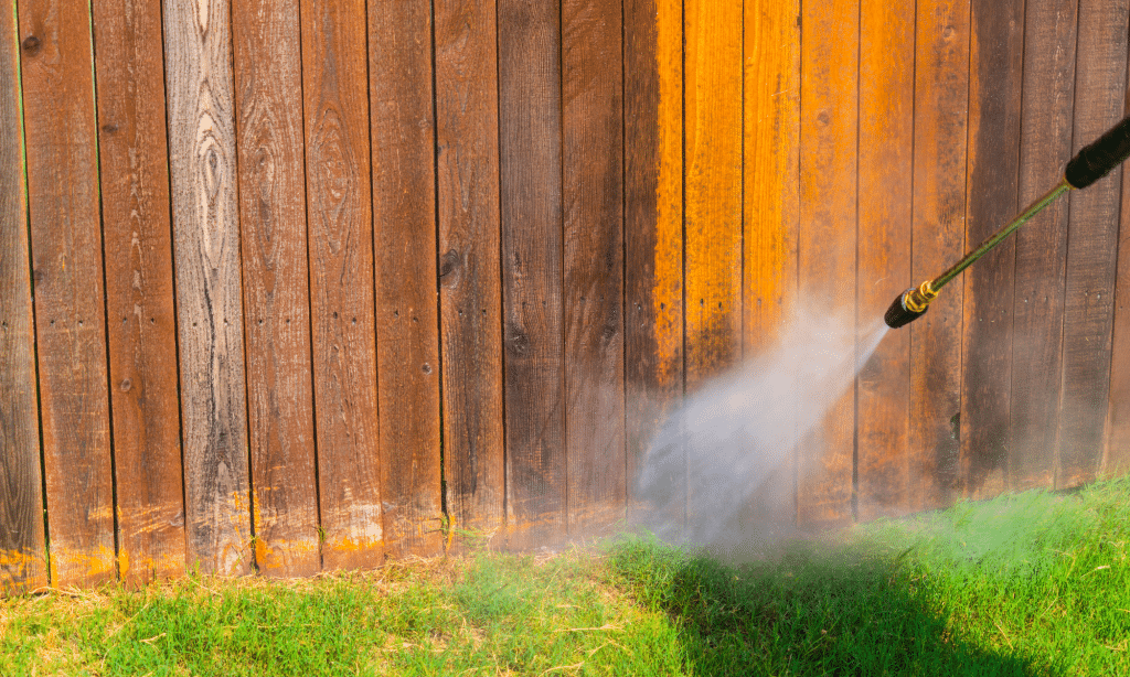 Water sprays from a pressure washer onto a wooden fence, revealing a clean section amidst the uncleaned areas